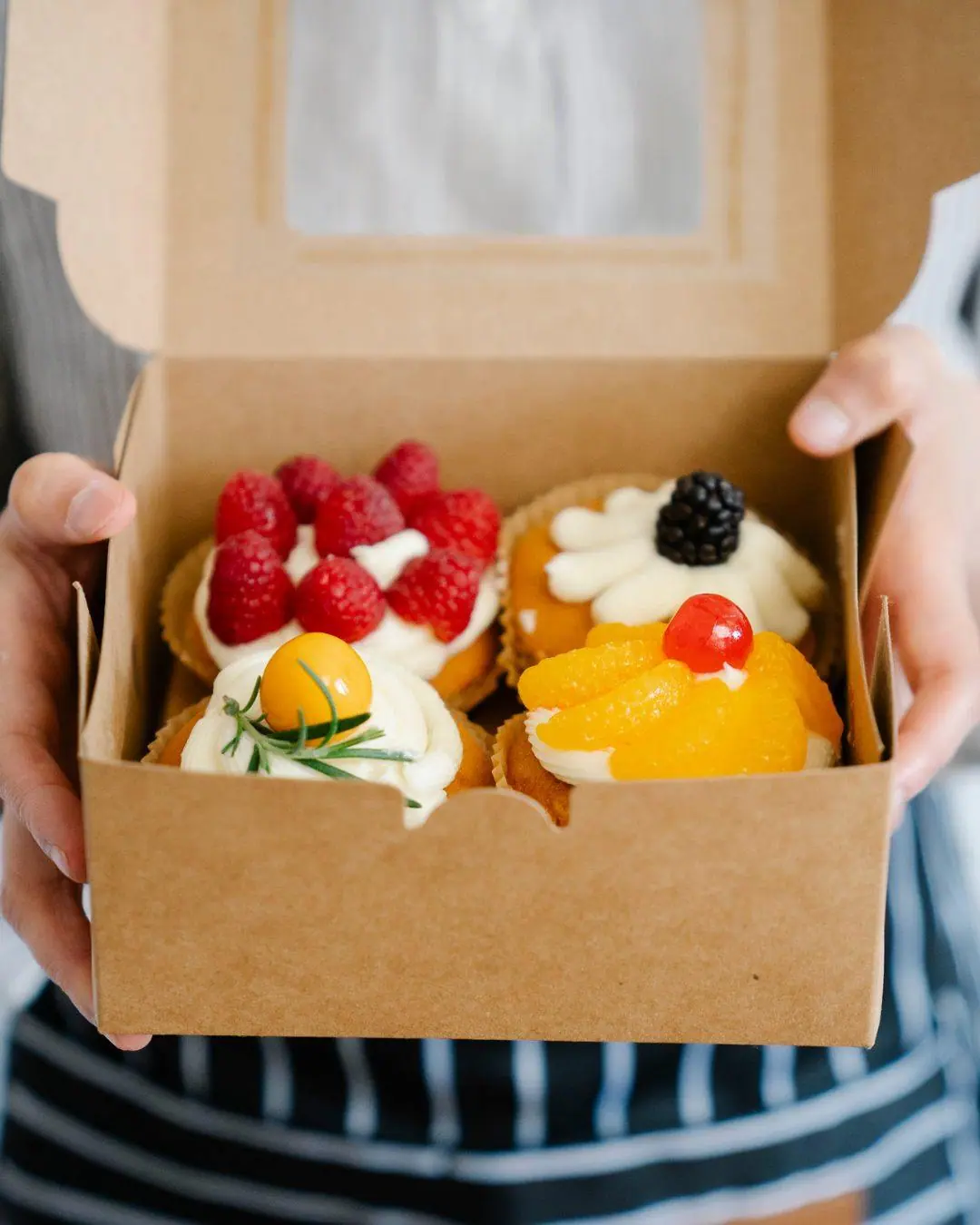 Four fruit-topped cupcakes in a kraft window box held in two hands